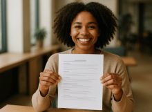 Uma pessoa sorridente, em ambiente interno iluminado suavemente, segura um currículo de forma confiante em um enquadramento médio e ângulo levemente lateral, transmitindo entusiasmo e expectativa profissional