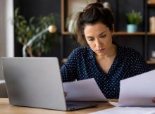 Uma mulher concentrada organiza documentos e revisa informações em um notebook, em um ambiente de trabalho moderno e bem iluminado